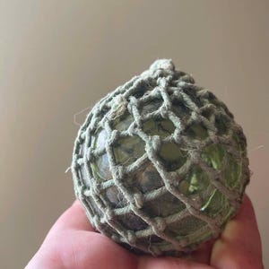 May include: A spherical, translucent light green glass float encased in a decorative, knotted beige jute rope netting. The glass orb features a smooth, rounded surface texture, while the surrounding twine creates a distinct grid pattern.