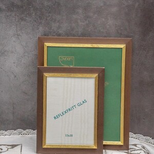 Może przedstawiać: Dwa brązowe ramki do zdjęć z pozłacanymi ozdobami. Mniejsza ramka ma napis "REFLEXFRITT GLAS" i "13x18" nadrukowany na passe-partout. Większa ramka ma napis "Jyden" nadrukowany na passe-partout.