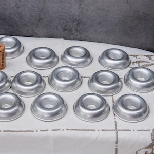 以下が含まれることがあります： 中央に穴の開いた銀色の円形モールドのコレクション。おそらくベーキング用。モールドは白い模様の布の上に配置され、背景にはコルクがあります。全体的なテーマは、キッチン用品またはベーキング用品を示唆しています。