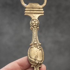 Puede incluir: Abridor de botellas de latón ornamentado con un mango decorativo. El abridor presenta una abertura en forma de corazón y un diseño ovalado en el mango. El latón tiene un aspecto vintage.