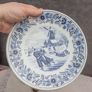 May include: A white ceramic plate with a blue and white Delft design. The plate features a winter scene with a windmill, a couple ice skating, and a sailboat. The border of the plate is decorated with blue and white floral designs.