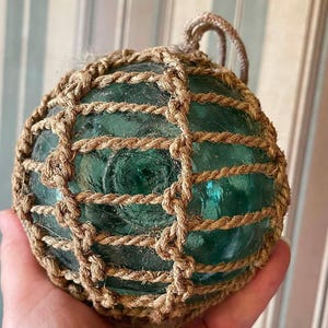 May include: A spherical, translucent sea-green glass float encased in a decorative, hand-knotted tan jute rope netting. The glass orb features a circular indentation at the center and includes a sturdy rope loop at the top for hanging purposes.
