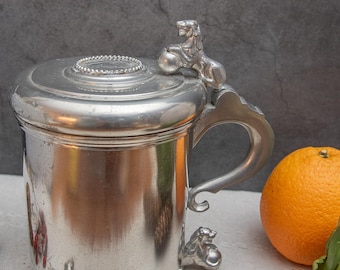 May include: A silver pewter tankard with a lid and a lion finial. The tankard has a handle and sits on three lion feet. The tankard is on a white surface with an orange and pine cones.