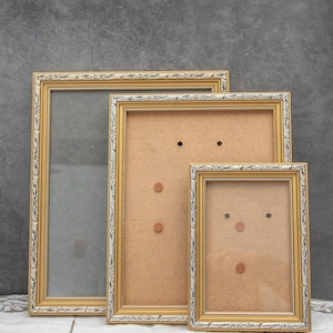 Könnte beinhalten: Drei goldfarbene Bilderrahmen in verschiedenen Größen. Die Rahmen haben eine verzierte Detaillierung und einen silbernen Akzent. Der größte Rahmen hat eine Glasfront, während die anderen beiden eine Kartonrückwand mit kleinen kreisförmigen Befestigungen haben.