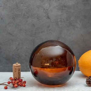 Puede incluir: Un flotador de vidrio grande, redondo y de color ámbar. El flotador está sobre una superficie blanca con un pequeño corcho y un racimo de bayas rojas.