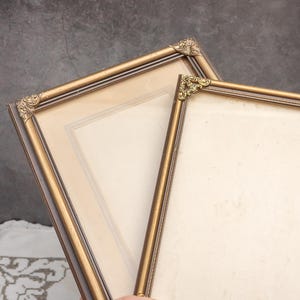 Può includere: Due cornici antiche color oro con dettagli angolari ornati. Le cornici hanno una forma rettangolare e sono tenute insieme. Lo sfondo è di colore grigio scuro.