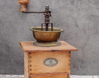 May include: An antique wooden coffee grinder with a brass bowl and handle. The grinder has a small drawer and a wooden knob. The design is vintage, with a rustic aesthetic. The grinder is set against a gray background.