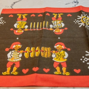 May include: A red and brown fabric kitchen towel with a repeating pattern of two figures holding objects. The figures are wearing red hats and yellow shirts. The objects are a stack of round brown items and a row of yellow sticks.