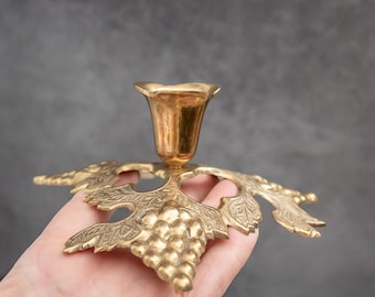 May include: A brass candle holder with a grape and leaf design. The candle holder has a cup-shaped top and a base shaped like a grape cluster with detailed leaves. The brass has a warm, golden color.