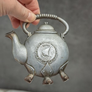 May include: A silver-coloured metal teapot with a rounded body, spout, and handle. The teapot has three small feet and a decorative emblem with a ship and the word "NORGE".