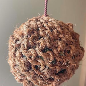 May include: A spherical object crafted from thick, brown, fibrous twine woven into a dense, textured knot pattern. The sphere is suspended by a single, thin, mauve-coloured braided cord attached to the top, creating a hanging decorative element.
