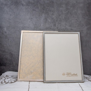 Könnte beinhalten: Zwei rechteckige Bilderrahmen mit silberfarbenen Metallrahmen. Einer hat einen hellbeigen Hintergrund, der andere einen weißen Hintergrund mit dem Text "Fotograf PP Lyshol". Die Rahmen stehen auf einer weißen Tischdecke. Geeignet für Fotos.