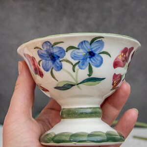 Op de afbeelding: Een witte keramische kom met een groene rand en een bloemenmotief. De kom heeft een voet en heeft blauwe, rode en groene bloemen.