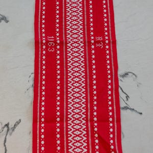May include: A red textile with a white geometric pattern. The design includes vertical stripes of white dots and a central diamond pattern. The numbers "1163" and "88" are visible on the textile.