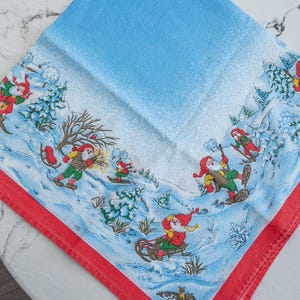 May include: A square, light blue Christmas-themed cloth napkin with a red border. The napkin features a winter scene with cartoon figures in red hats and green outfits skiing, sledding, and snowboarding in a snowy landscape.