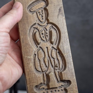 May include: A wooden cookie cutter in the shape of a person wearing a hat and pants. The cutter has a detailed design and is made of light brown wood.