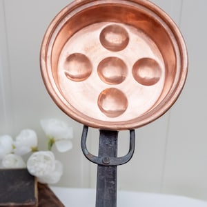 Puede incluir: Una poachadora de huevos de cobre con cinco hendiduras circulares. La sartén tiene un borde redondeado y un mango de metal oscuro. El mango está unido a la sartén con una junta remachada. Es un utensilio de cocina.