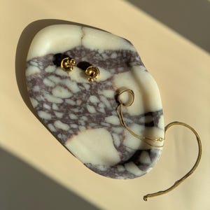 May include: A white marble tray with brown and purple marbling. The tray is holding a gold chain necklace, a gold ring, and a pair of gold earrings.