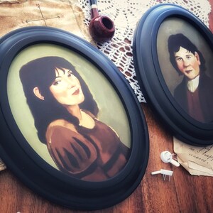 May include: Two oval-framed portraits with black frames. One portrait features a woman with dark hair and a brown dress. The other portrait shows a man in a dark coat and white shirt. A pipe and paper are in the background.