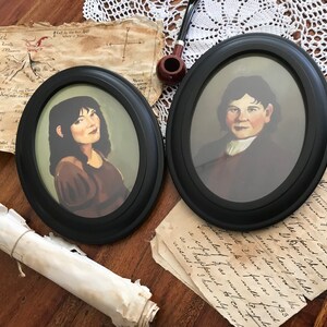 May include: Two oval framed portraits with black frames. One portrait depicts a woman with dark hair and a brown dress. The other portrait shows a man in a dark coat and white shirt. The portraits are on a wooden surface with antique paper and a pipe.
