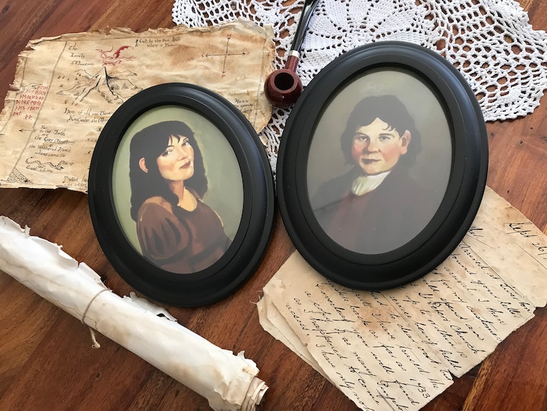 May include: Two oval framed portraits with black frames. One portrait depicts a woman with dark hair and a brown dress. The other portrait shows a man in a dark coat and white shirt. The portraits are on a wooden surface with antique paper and a pipe.