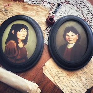 May include: Two oval portraits in black frames. One depicts a woman in a brown dress, the other a man in a dark coat. The portraits rest on a wooden surface with aged paper and a pipe.