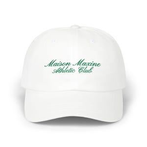May include: White baseball cap with a curved brim. The cap features the embroidered text "Maison Maxine Athletic Club" in green script across the front. The hat is a classic style, suitable for casual wear.