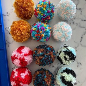May include: Ten pairs of colorful pom-pom earrings. The earrings are made of yarn and have a variety of colors and patterns, including blue, green, red, white, and black.