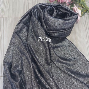 May include: A black sparkly scarf with a fringed edge. The scarf is folded and draped over a wooden surface. The word "Pirilty" is printed on the scarf.