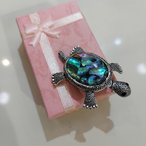 Puede incluir: Un broche de tortuga de plata con una incrustación de nácar abalone verde, azul y morado. El broche está en una caja de regalo rosa con una cinta blanca.