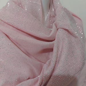 Puede incluir: Una bufanda rosa claro adornada con brillantes plateados. La bufanda está drapeada sobre una cabeza de maniquí blanca, mostrando su textura suave y detalles brillantes. La tela está doblada, creando profundidad y resaltando el brillo disperso.