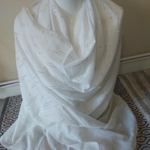 May include: A white scarf featuring a subtle pattern of thin, horizontal silver metallic lines and small dots. The fabric has a lightweight, textured appearance, and the edges are finished with a delicate, scalloped white lace trim detail.