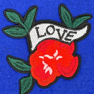 May include: Embroidered iron-on patch featuring a red flower with green leaves and a white banner with the word "LOVE" in black lettering.
