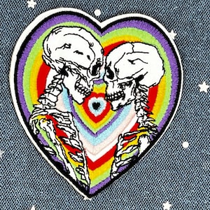 May include: Embroidered iron-on patch featuring two skeletons embracing inside a rainbow heart. The skeletons are white with black outlines and the heart is filled with concentric circles of rainbow colors.