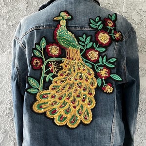 May include: A denim jacket with a large, colorful peacock and floral sequin patch on the back. The peacock features green, gold, and red sequins, with a detailed tail and a floral design.