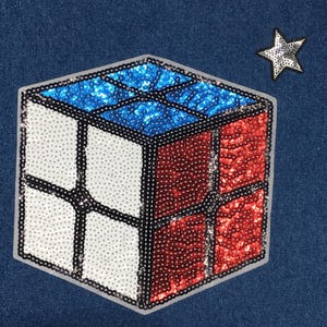 May include: A red, white, and blue sequined Rubik's Cube patch. The patch is shaped like a cube and has a star in the upper right corner.