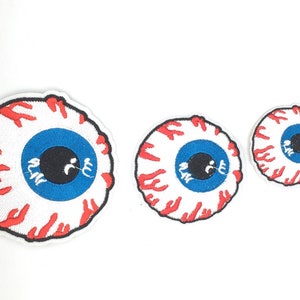 May include: Three embroidered eye patches with a white background. Each patch features a blue iris, a black pupil, red veins, and a white sclera. The patches vary in size, with the largest on the left and the smallest on the right.