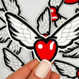 May include: Embroidered patch featuring a red heart with white wings outlined in black. The wings have detailed white feathering. The patch is held in a hand against a background of similar patches.