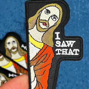 May include: Embroidered patch featuring a stylized image of Jesus with long hair and a beard, wearing a red and orange robe. The patch has the text "I SAW THAT" in white letters on a black background. Several similar patches are visible.