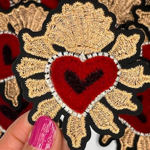 May include: A close-up of a red velvet heart with a black outline, surrounded by a gold embroidered halo. The heart is a religious symbol and is often used in crafts and decorations.