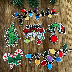 May include: A collection of Christmas-themed patches on a wooden surface. The patches include a Christmas tree, candy canes, string lights, and a hand holding an ornament. The text "season's greetings" is also featured.