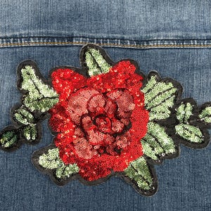 May include: A denim jacket with a large, sequined red rose applique. The rose is the focal point, with green sequined leaves and a black outline. The jacket's blue denim provides a contrasting background.