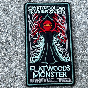 May include: A black embroidered patch with the text "Cryptozoology Tracking Society" and "Flatwoods Monster" with a red and green illustration of a creature with large eyes.