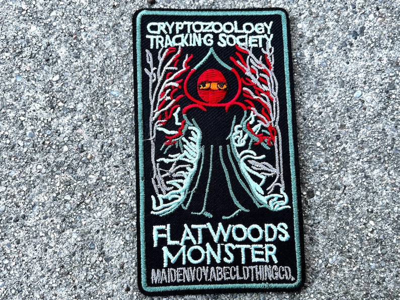 May include: A black embroidered patch with the text "Cryptozoology Tracking Society" and "Flatwoods Monster" with a red and green illustration of a creature with large eyes.