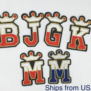 May include: Embroidered patches featuring the letters B, J, G, K, and two M's, each topped with a crown. The letters are in red, white, and blue, outlined in gold, and set against a black background.