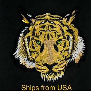 May include: Embroidered patch of a tiger's face with green eyes and a yellow and white striped pattern. The tiger's face is facing forward and the background is black.