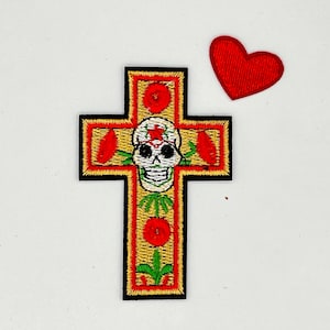 May include: An embroidered iron-on patch featuring a yellow cross with a red border. The cross is decorated with red flowers and a green stem. In the center of the cross is a white skull with black eyes and a red nose.
