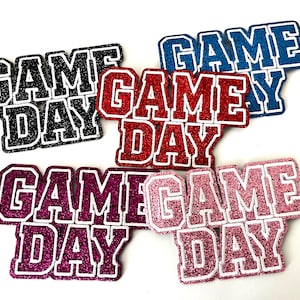 May include: A collection of glittery "Game Day" cutouts in various colors, including black, red, blue, purple, and pink. Each cutout has a white outline, creating a bold, eye-catching design. Perfect for sports-themed crafts and decorations.