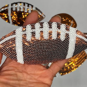 May include: Close-up of several football-shaped embellishments. The footballs are brown with white stitching and are covered in sequins. The embellishments are held in a hand against a white background. The sequins shimmer.