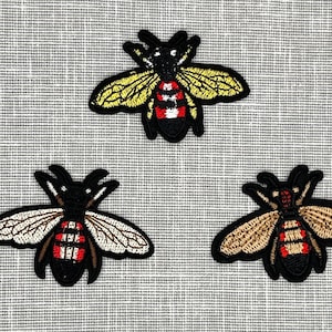 May include: Three embroidered bee patches in different colors. The bees are black with red and white stripes and have yellow, white, or brown wings.